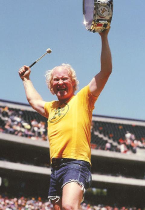 man in cutoff shorts and tee shirt with arms upraised, holding a drum and baton