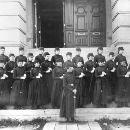 three lines of young women dressed in dark skirted military uniforms and caps, holding with swords or rifles, looking left while posed on the steps leading into a large building. An inswet at the upper left is a portrait of a man with a moustache