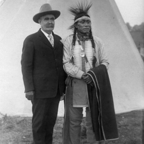 Caucasian man with big moustache, in cowboy hat, suit and tie, stands next to Arapaho man in traditional headband, chest plate and breeches, with a blanket over one arm