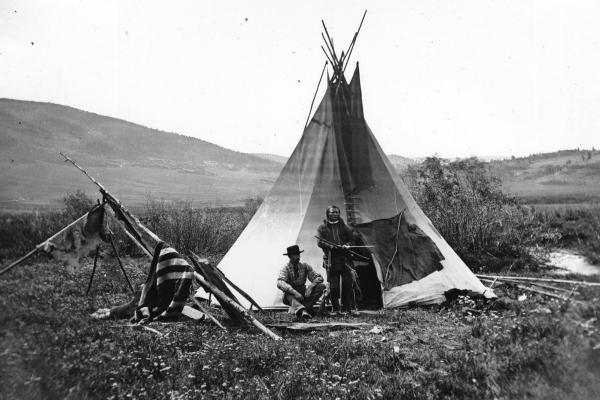black and white landscape of open plains and a creek with a teepee. Ferdinand V. Hayden and an unidentified American Indian sit and stand in front, respectively.