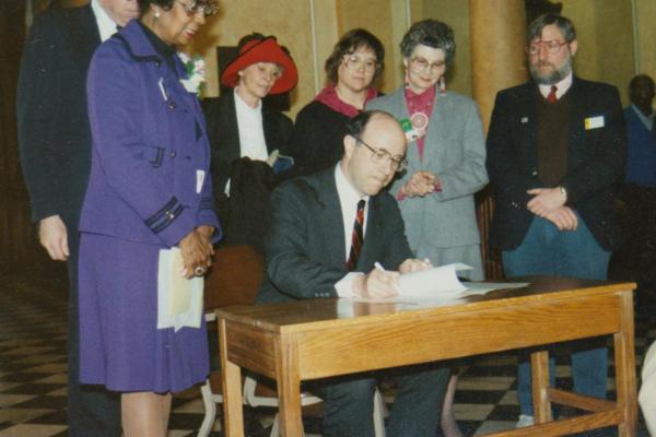 A group of women and men stand around a desk where a man is signing a document