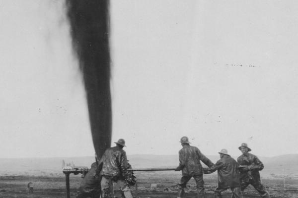 Five men around a plume of oil rocketing into the sky; they are grasping a very large valve