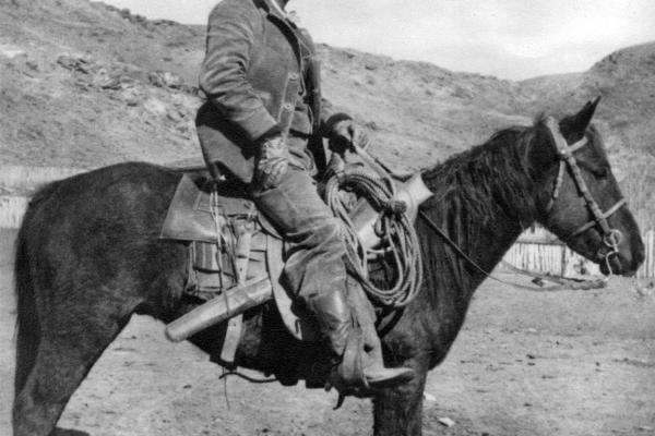 a man wearing a cowboy has, chaps, boots, and gloves, on a saddled horse, in profile