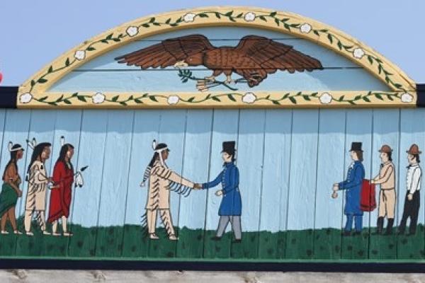 painted mural of two groups of three men: on the left, Native American, on the right Caucasians; both groups look to the center where a Native shakes hands with a Caucasian. Above this scene is painted an eagle with olive branch in one claw and arrows in another