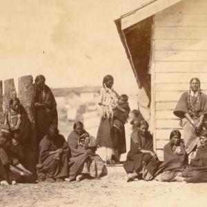 Lakota Sioux women, children and, against the right-hand end of the fence, a man, possibly the Oglala Lakota, High Wolf, at the Indian agent's quarters at Fort Laramie, 1868. Sherman collection of Gardner photographs; NAMI.