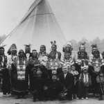 A large group of Northern Arapaho traveled to London and Paris in 1923-24 to appear in live performances staged before showings of the new movie "The Covered Wagon."  Few of the photos show women and children as well as the men; here, the group poses in a park in London. Ed Farlow kneels front and center, Yellow Horse stands at far right and Jack Shavehead stands third from right. The rest are unidentified. American Heritage Center.