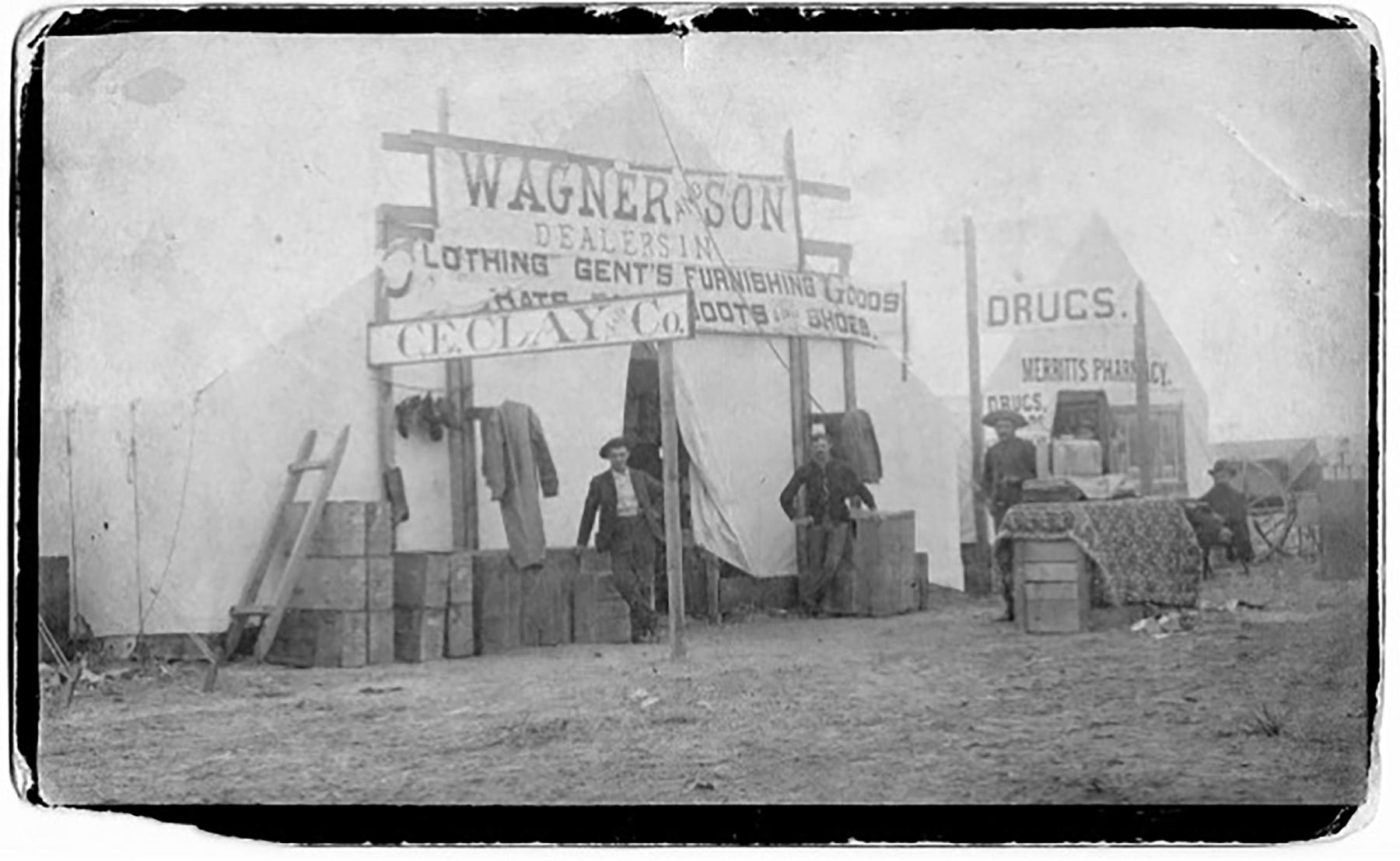 When Zindel first arrived, Douglas was a tent city, as seen in this 1886 photograph. Zindel was credited with building the first business and house after the city was plated. Large Canvas tent with signs: "Wagner and Son: Clothing, Gent's Furnishings, Goods, Hats, Boots, Shoes," "C.E.Clay & Co.," "Drugs, Merritt's Pharmacy." Four men pose in front, along piles of wood boxes