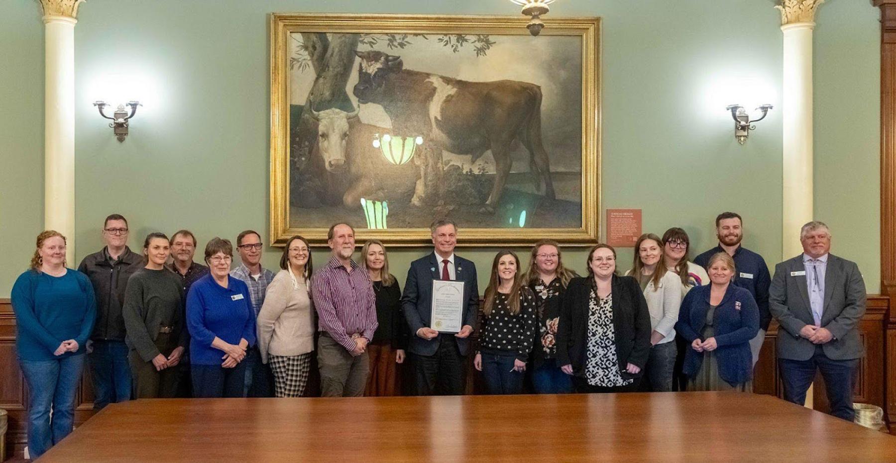 Governor’s Proclamation 2025, May 6, 2025. Photo by Denise Hawkins. A group of citizens stands to left and right of Wyoming Governor Mark Gordan, who is holding a proclamation and smiling. There is a large oil painting of a bull on display behind them.