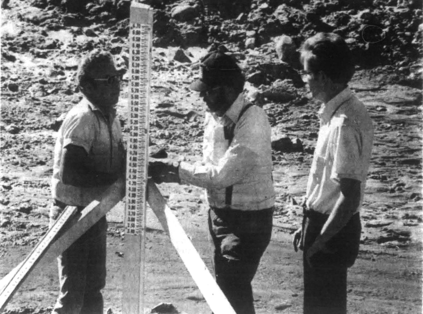 U.S. Geological Survey hydrologic technician Joe Wallace (left), hydrologist Jim Rankl (center), and assistant city engineer David Benner install a flood water staff gauge at Windmill and Sheridan on Dry Creek in October 1986. Three men observe a flood gauge along a creek
