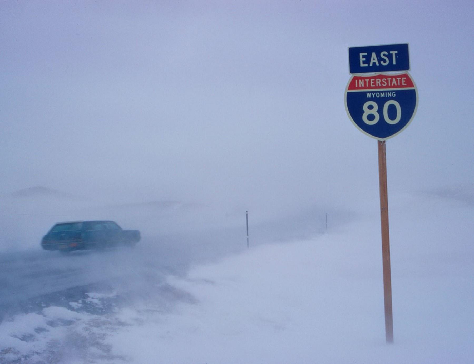 Conditions like this on Interstate 80 between Laramie and Walcott Junction led to the infamous name for this stretch of highway as the “Snow Chi Minh Trail.” Interstate 80 near Arlington, December 1972. Wind- and snowswept road with a vehicle in the spindrift, and a sign for "EAST - Interstate 80" on the side of the highway.