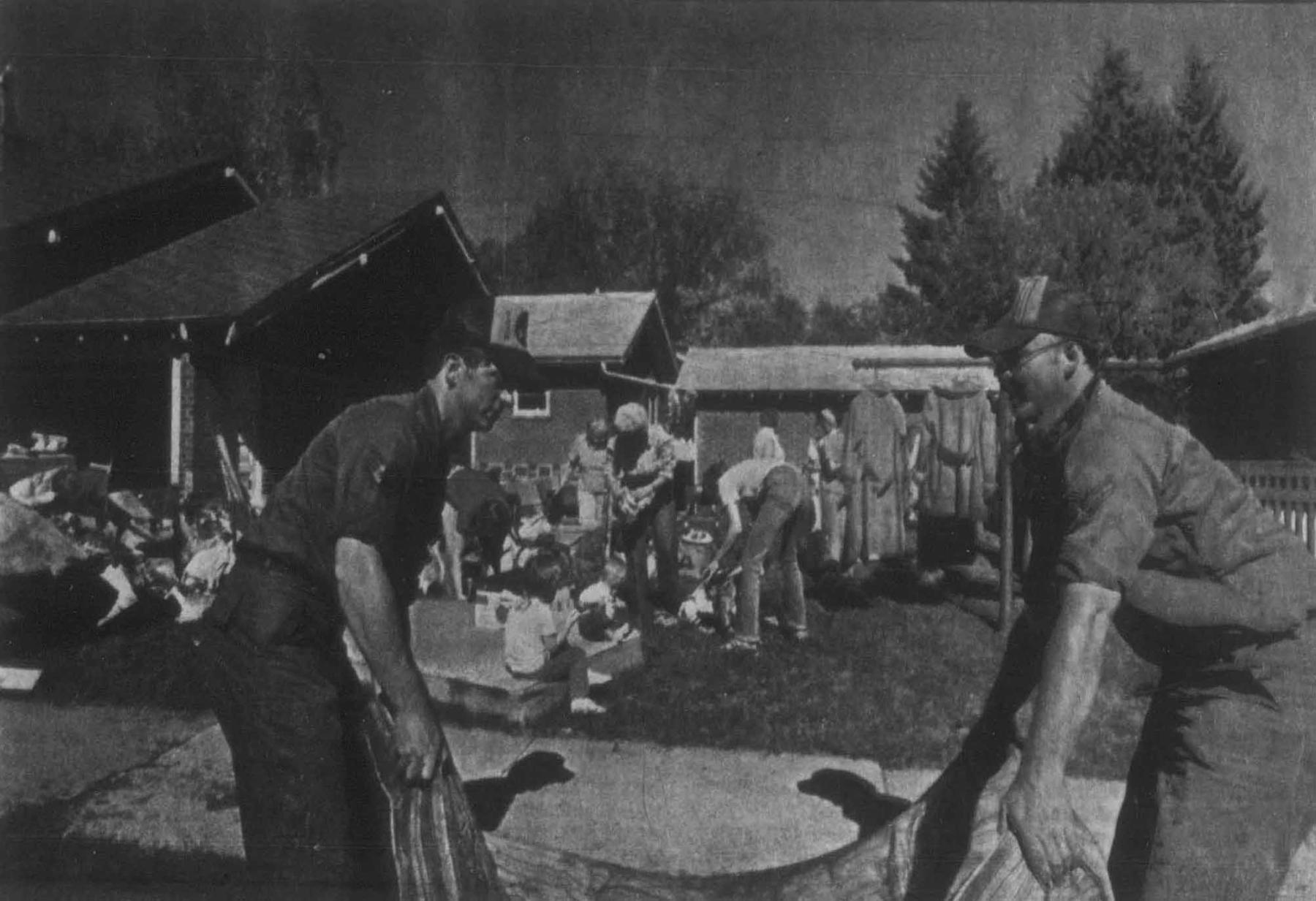 Two men in the foreground hold a tarp while a group of other peole are gathered in the bckground sorting through household articles outdoors