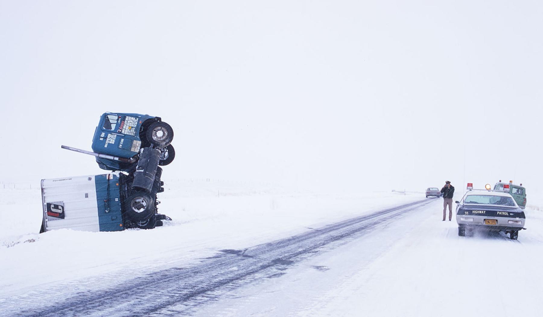 Interstate 80 near Arlington, December 1972. Semi-trailer jack-knifed and on its side next to an icy highway in whiteout conditions. A state trooper is parked opposite, standing outside his patrol car observing.