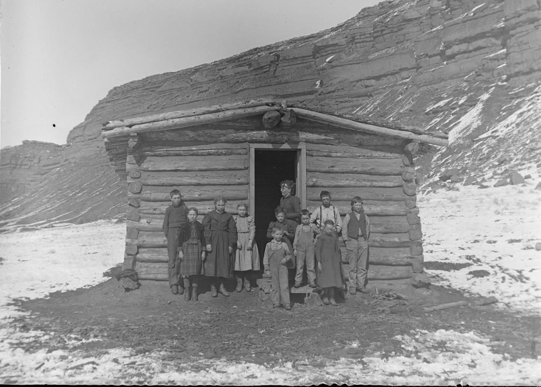 black and white photo of a one-room log building with ten children and a women standing out front. 