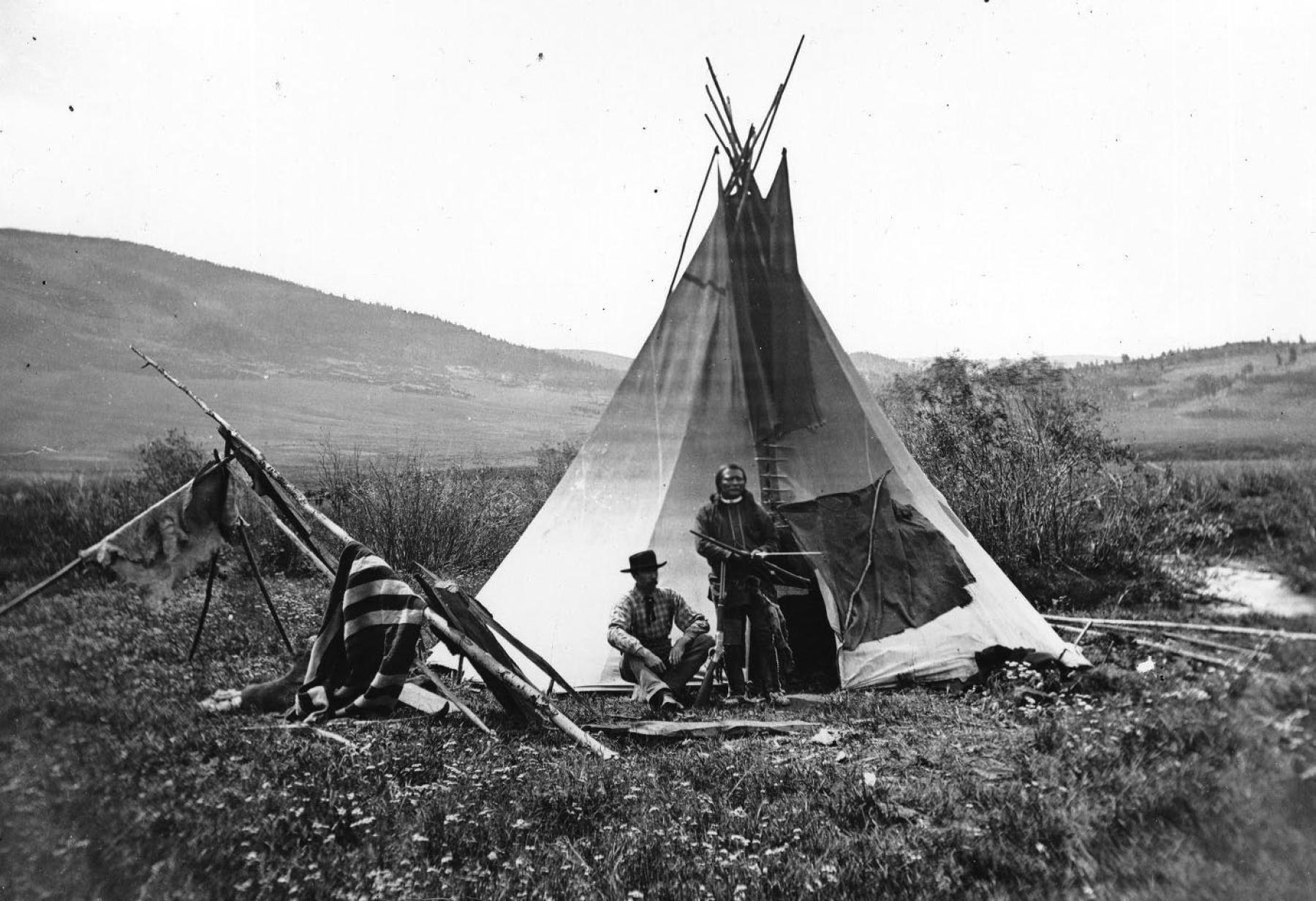 black and white landscape of open plains and a creek with a teepee. Ferdinand V. Hayden and an unidentified American Indian sit and stand in front, respectively.