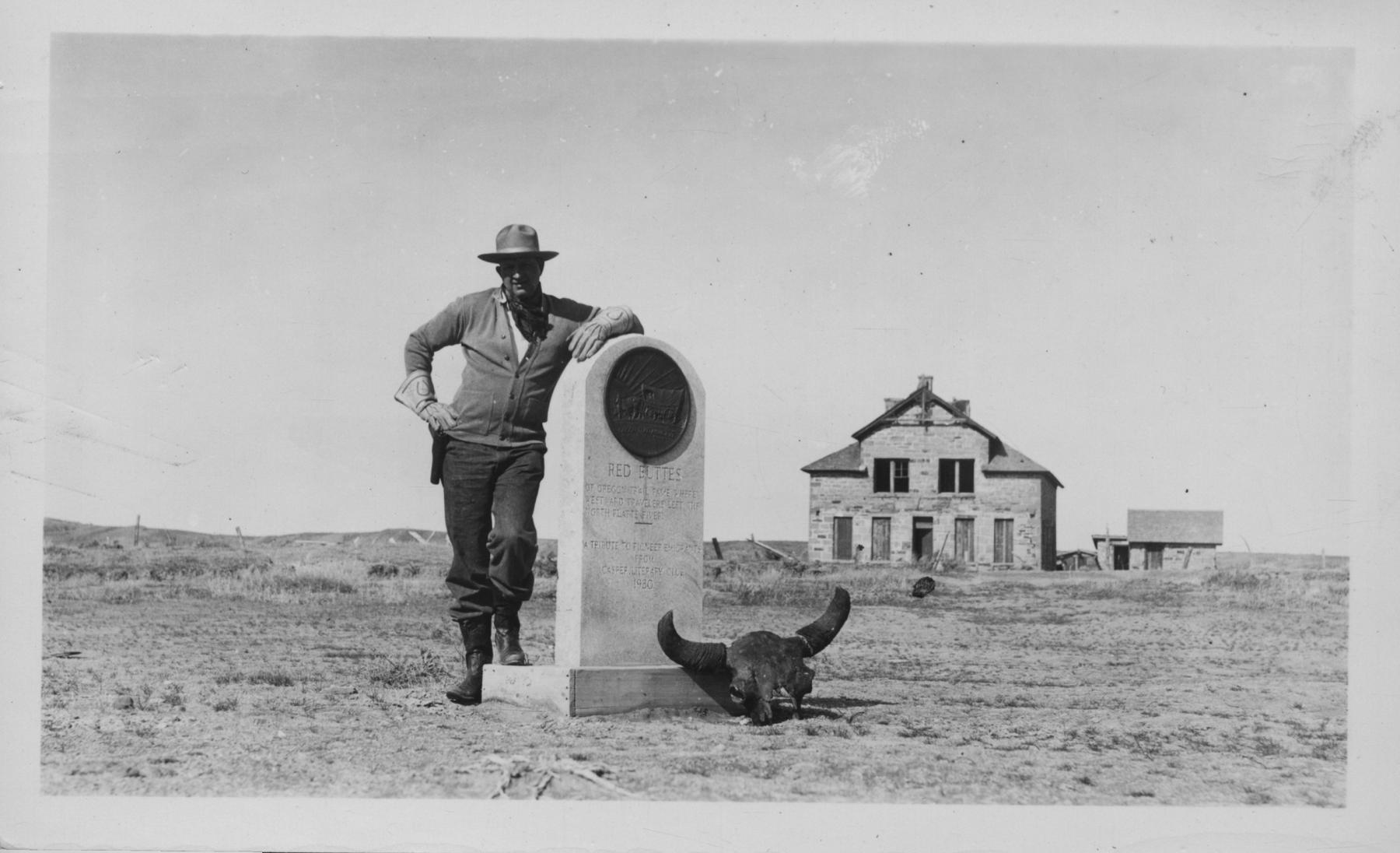 A man in a cowboy hat with holstrered gunbelt leans on a small monument with a bison skull at its feet. A two-story stone house is in th background, set on a wide open plain.