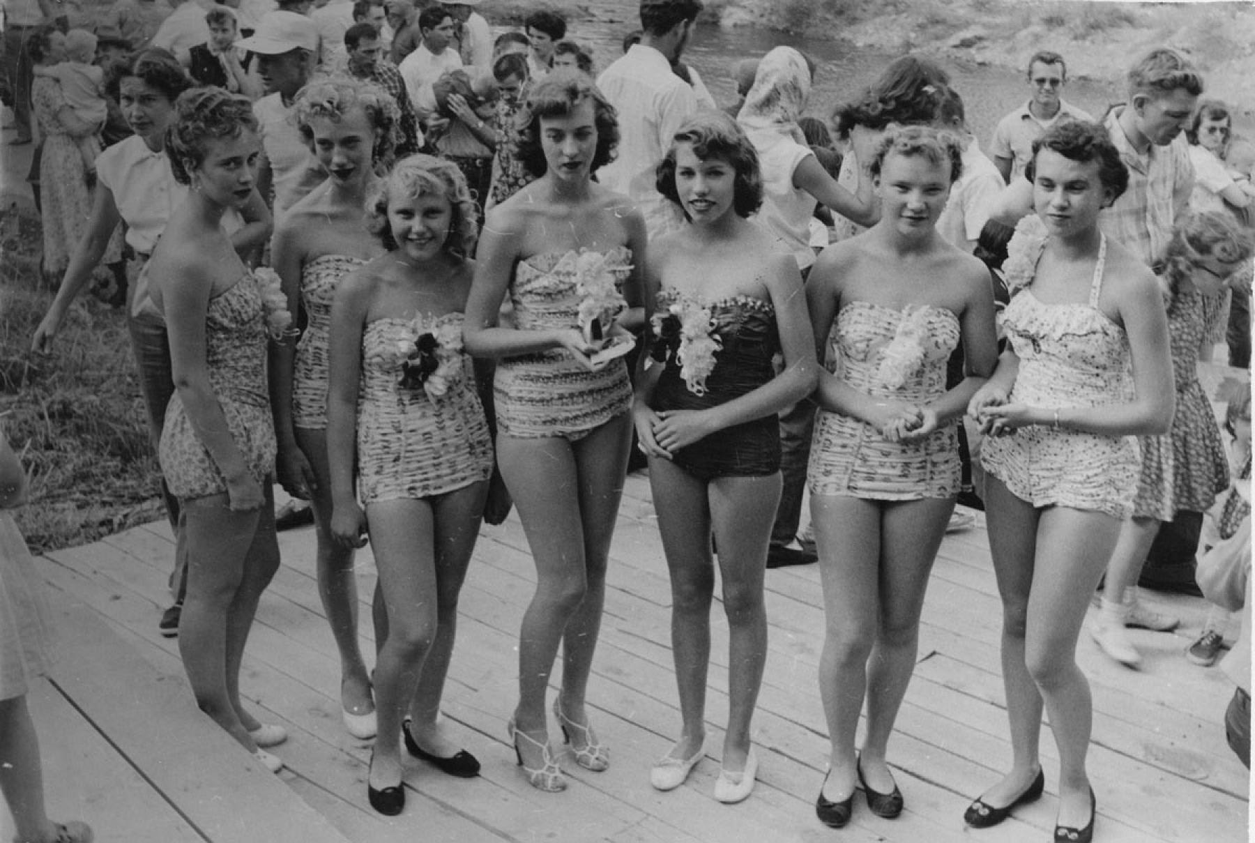 Seven women in one-piece bathing suits pose, standing in a row