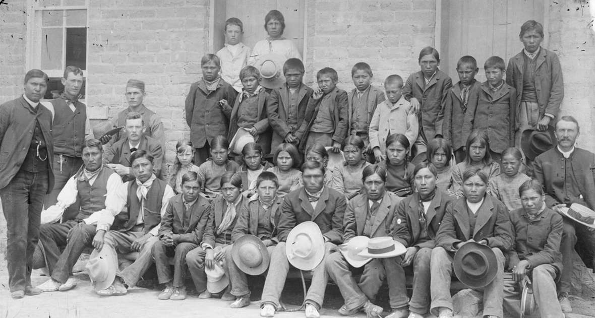In 1883, the Episcopal priest Dr. John Roberts, seated at right with hat on his knee, founded the first mission school on the reservation. Later he was joined by the Rev. Sherman Coolidge, standing at far left, an Arpaho who had been raised in the family of a white Army officer and educated in the East. This photo of boys in the school may date from around 1890. Note that the boys are all wearing moccasins. American Heritage Center