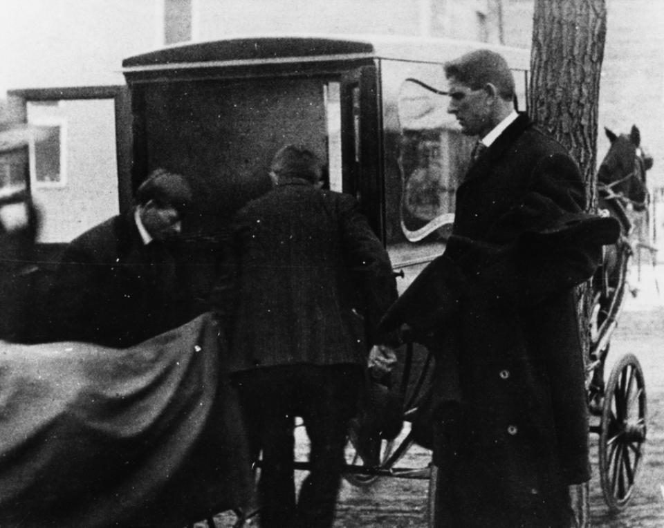 Men in dark clothing move a shrouded body into a horse-drawn carriage as a third man looks on.