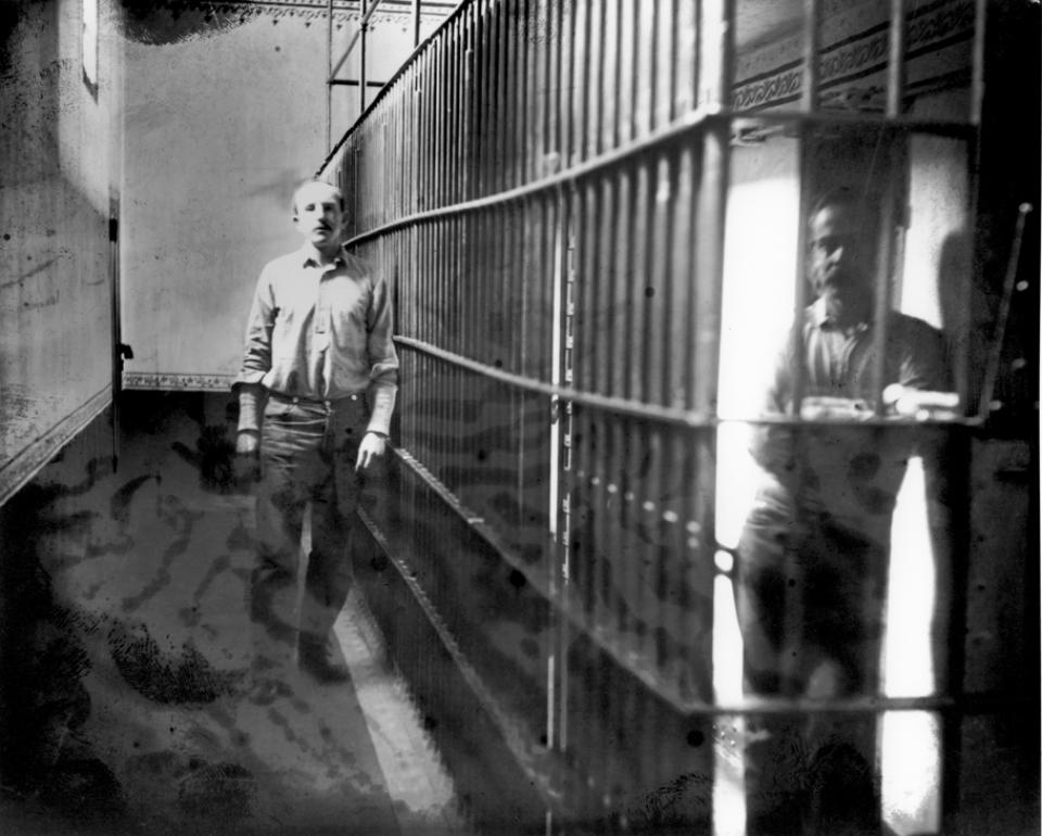 This eerie image of Tom Horn in his jail cell was taken while he awaited his execution. Photo courtesy of the Wyoming State Archives. Tom HOrn standing by cell bars, looking at the camera, while another person looks out from another cell
