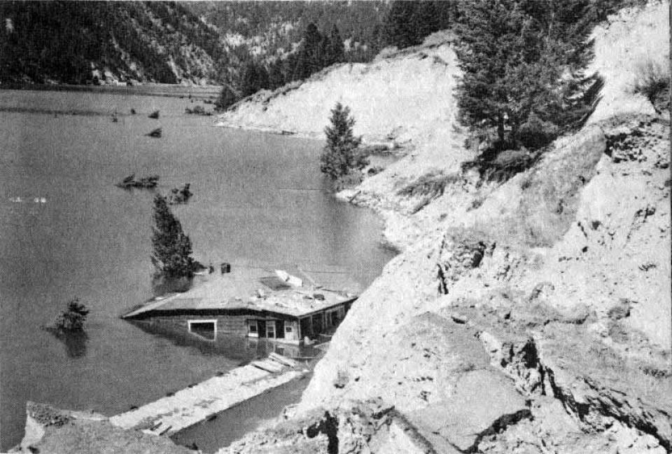 The edge of a lake whener a cabin tilts into the water, which rises to its roofline on one side