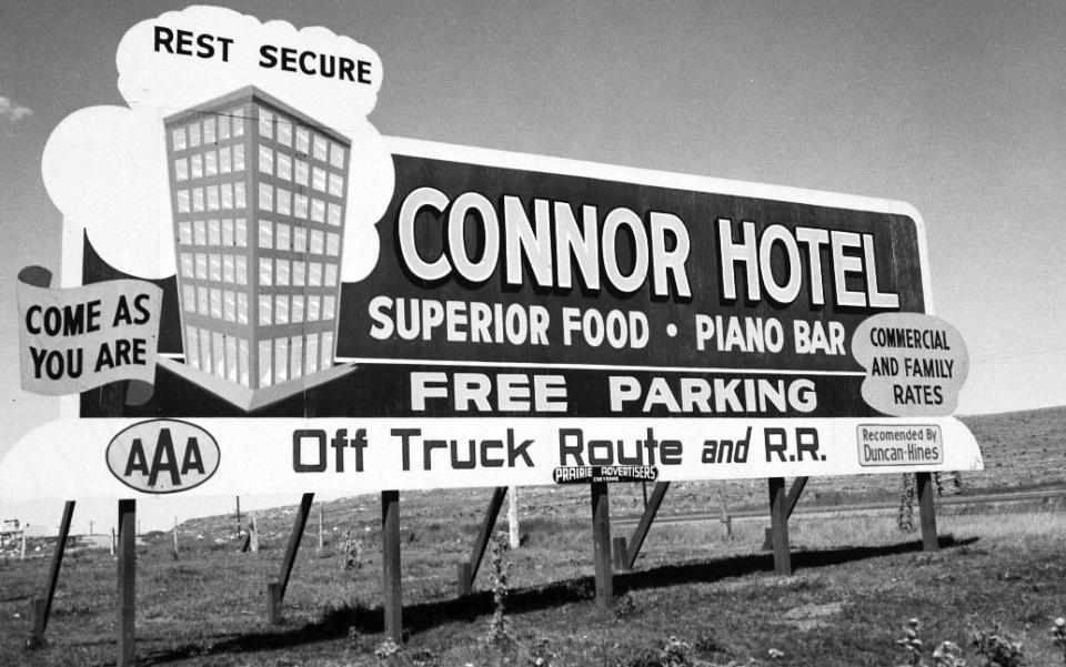 By 1965, billboards—like this one near Laramie—were seen as eyesores, leading the federal government to control their placement along highways. Billboard reads: "Connor Hotel / Superior Food / Piano Bar / Free parking / Off truck route and RR / Come as you are / Rest secure." There is a triple A logo and an image of a multi-story rectangular building.