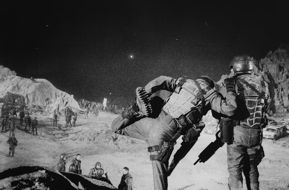 two costumed soldier-actors look over the landscape; one is stretching his left leg while the other holds him steady