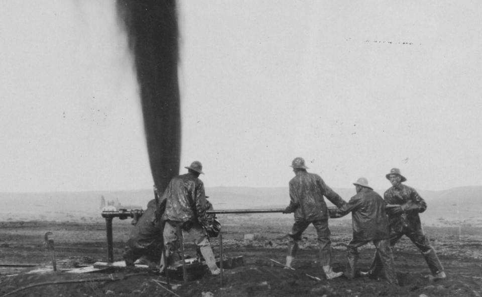 five men surround a jetting fountain of oil while attempting to turn the enormous valve on the drill hole