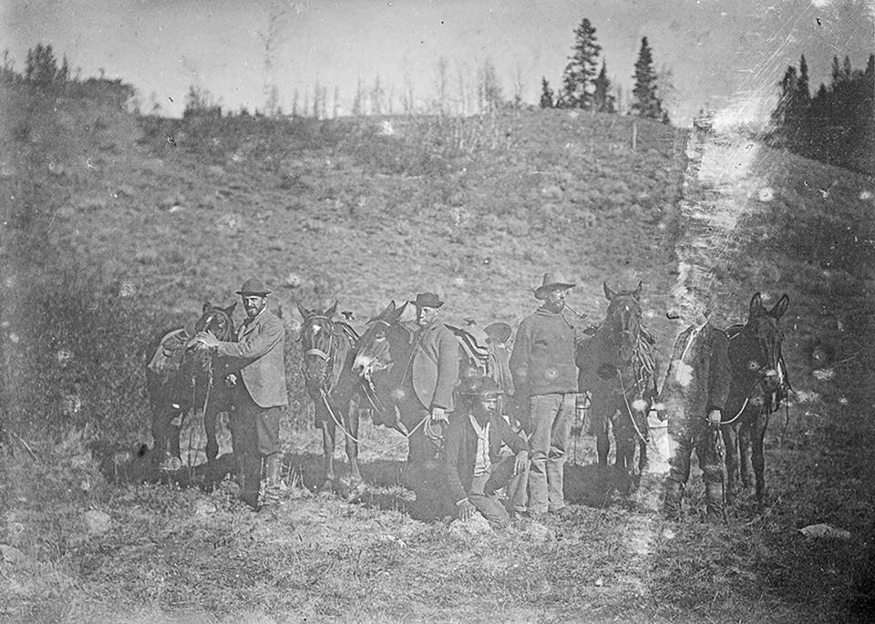 five men stand next to their horses and mules in an open, scrubby area