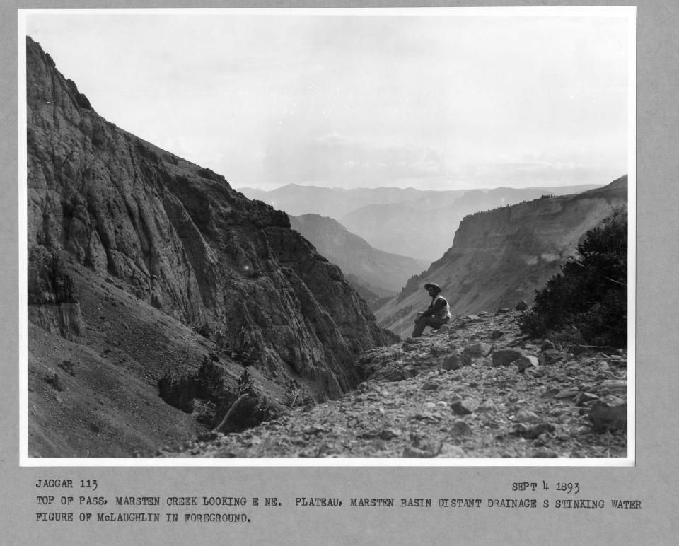 black and white photo looking down a steep valley, with a man seated on the rim