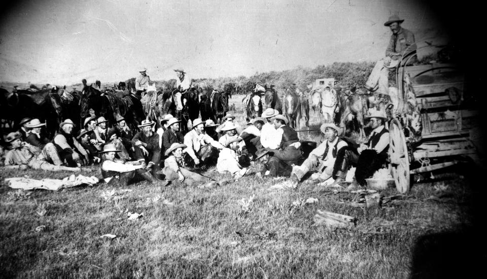 seated and mountede cowboys pose in a larg e group, including several women in dresses, and a large wagon