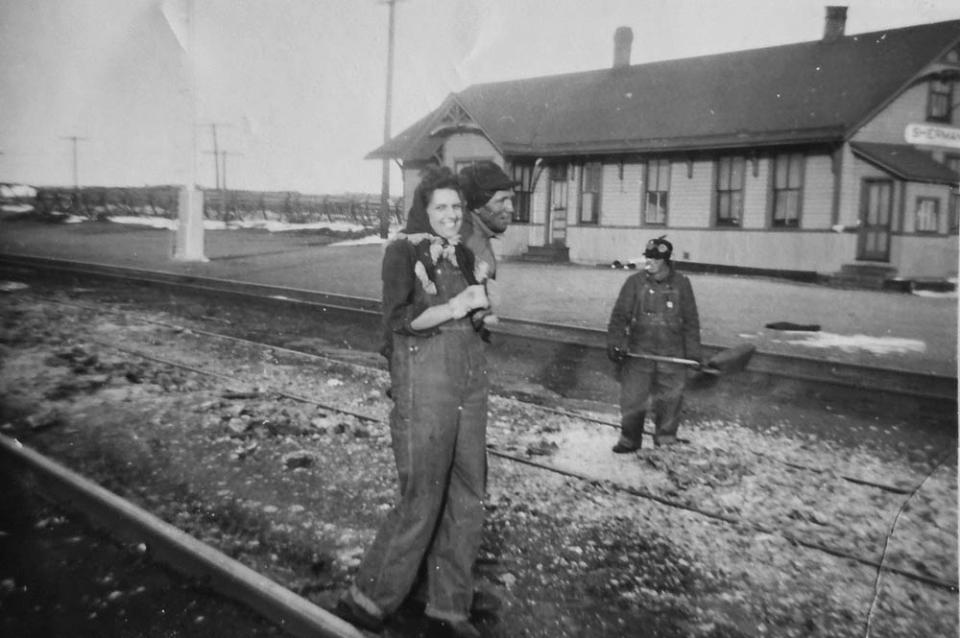 woman in overalls stands near railroad tracks with two men in work clothes; a train depot is in the background