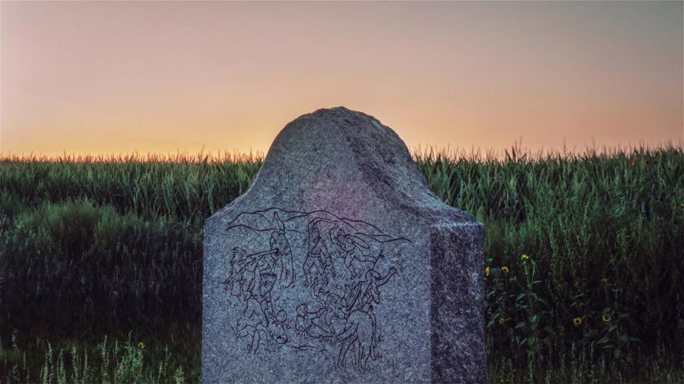 the upper half of a stone memorial marker, etched with figures fighting, against a background of tall grasses and morning light
