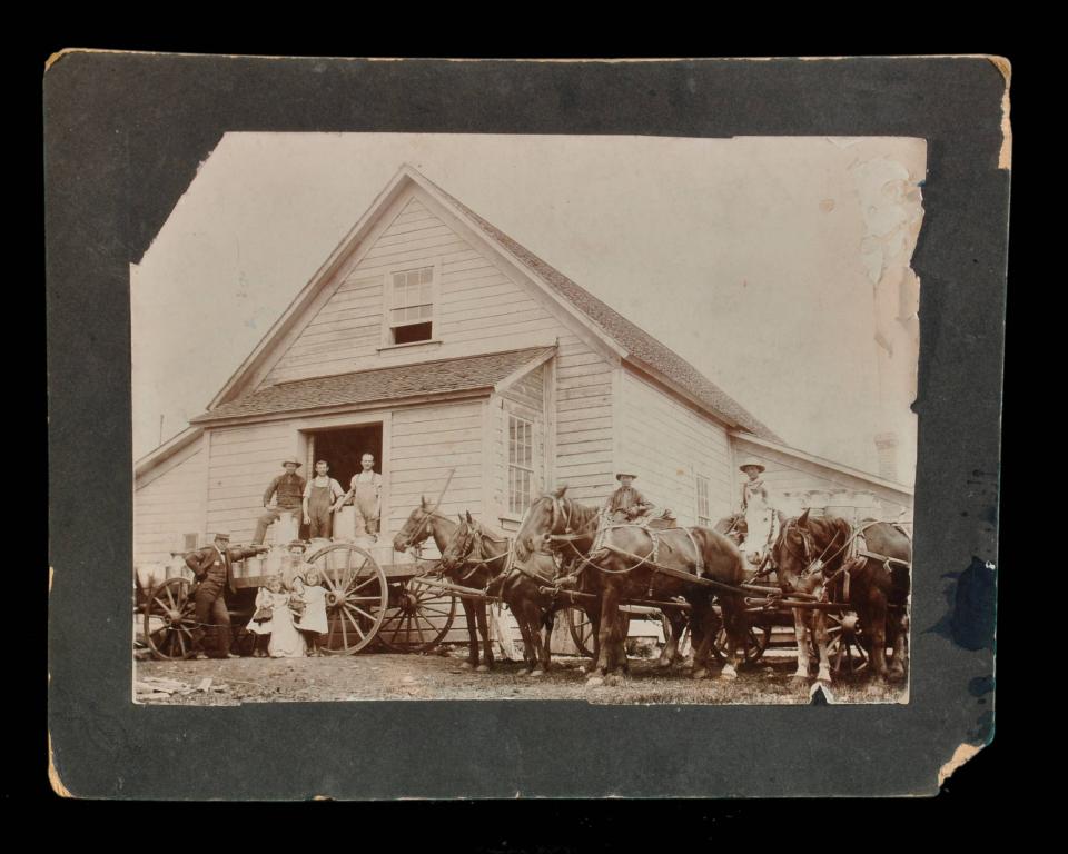 Men and women pose standing on and by a wagon loaded with milk cannisters, attached to a team of horses outside a building witht peaked roof