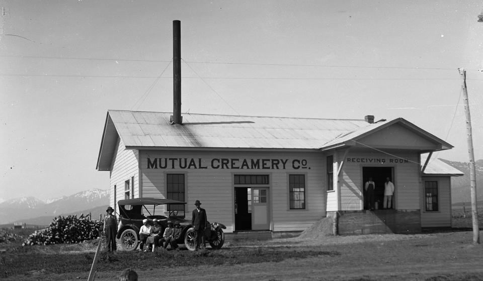 Men pose seated on the running board of an early automobile, parked in front of a building painted with the sign "Mutaul Creamery Co"