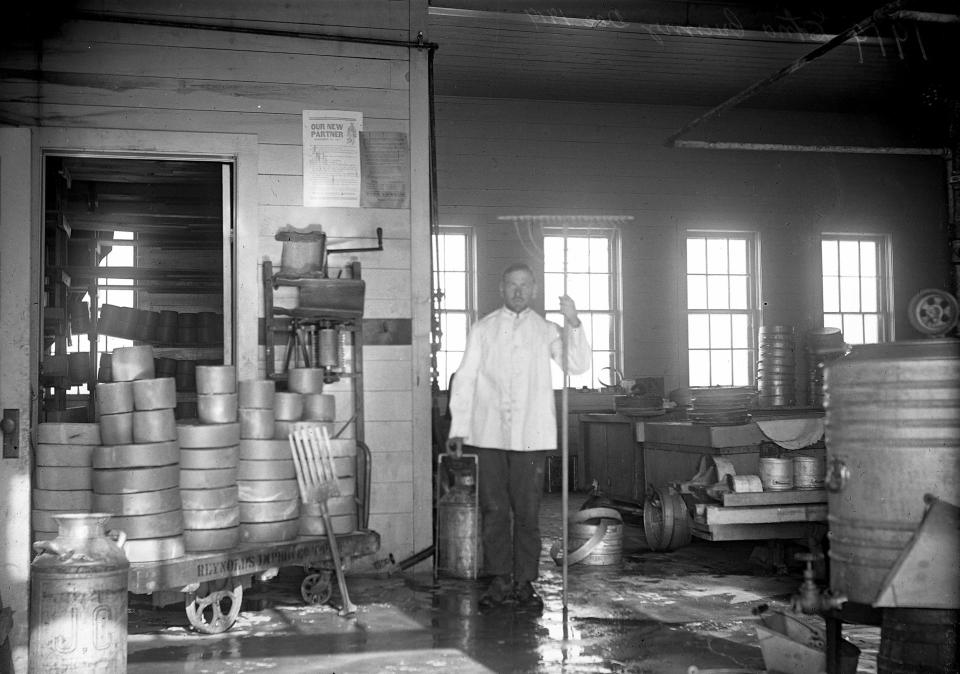 A man in a white jacket poses amidst various dairy and cheese processing equipment in a windowed room