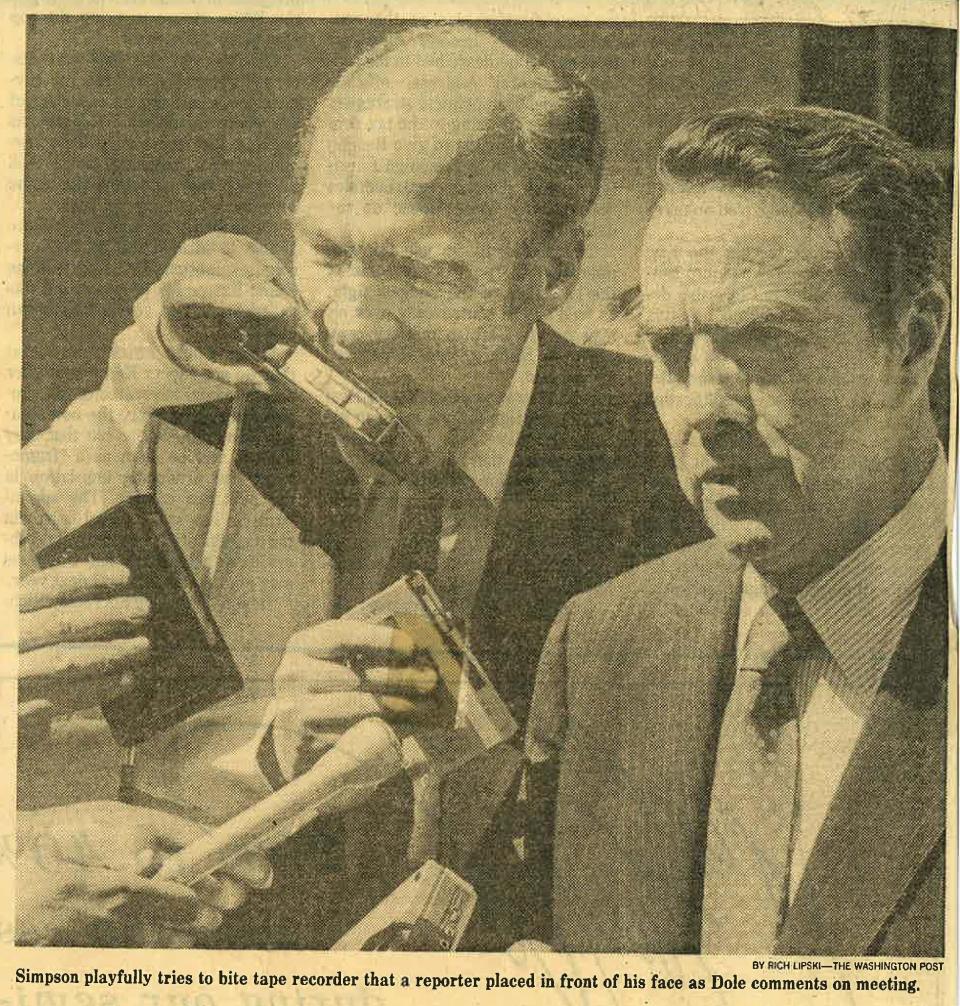 Newspaper clipping showing Alan Simpson biting at a hand-held tape recorder while standing behind Bob Dole