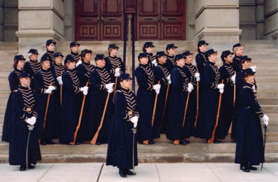 Rows of women wearing military skirted uniforms and caps, holding rifles and all looking left, on the steps of a large building
