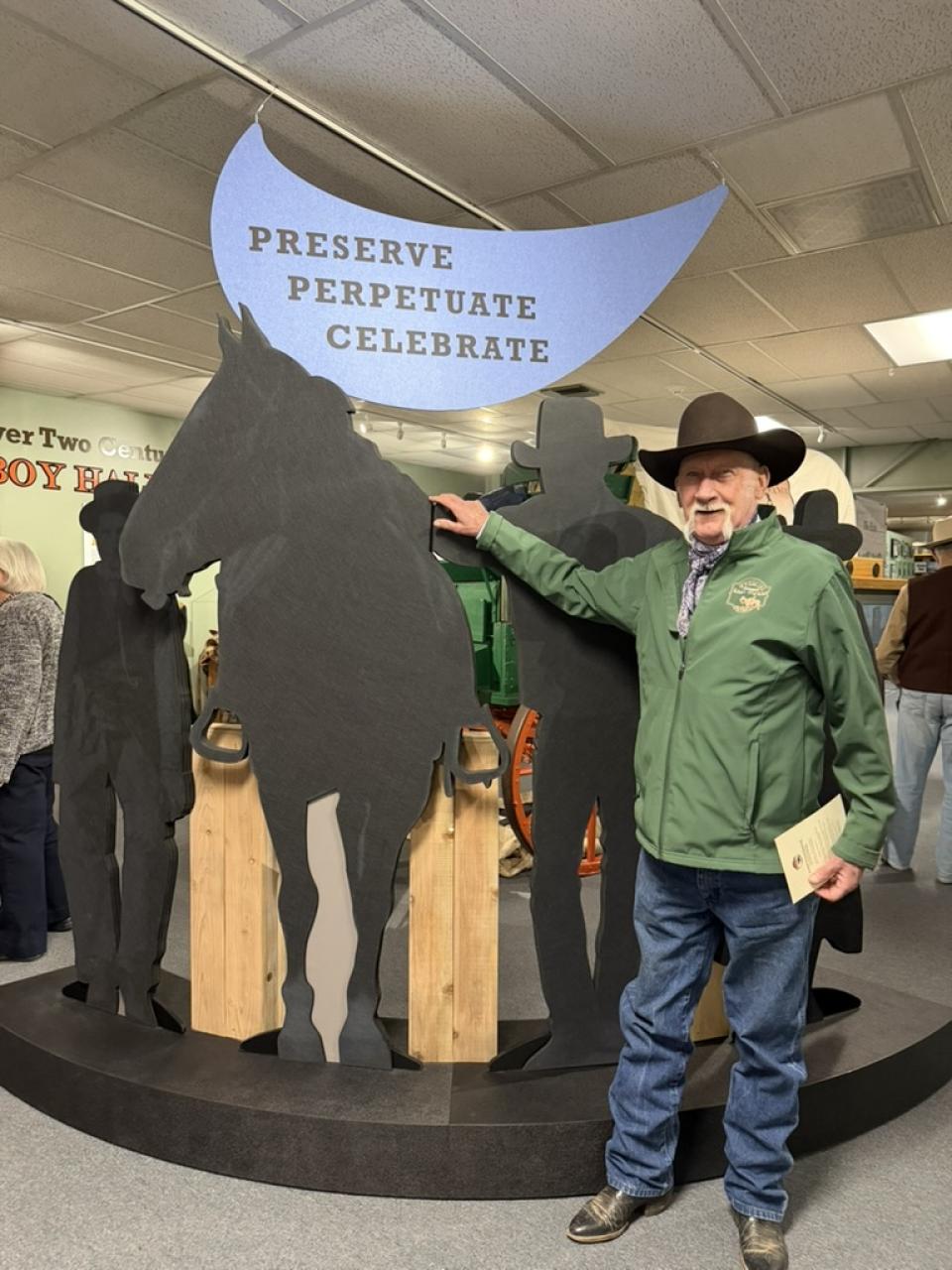 A man in a cowboy hat has his hand on the cut out of a horse, under the "Preserve Perpetuate Celebrate" sign