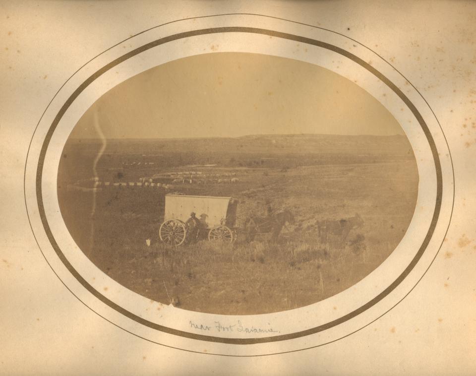 sepia toned phot in an oval mat showing a man standing on the side of a tall, mule-drawn wagon, with a covered wagon train winding in the distant plain