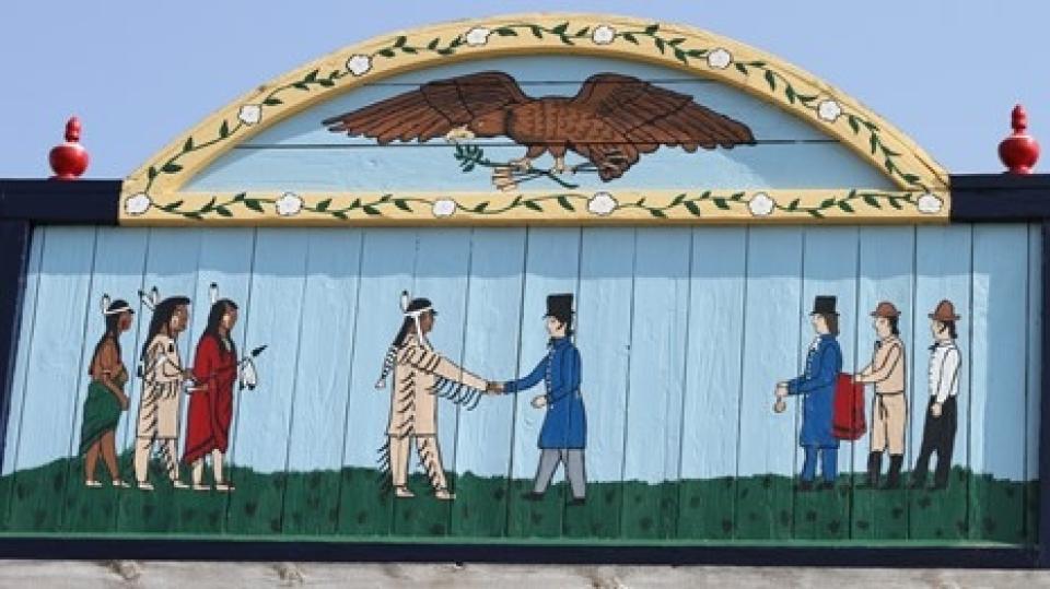 painted mural of two groups of three men: on the left, Native American, on the right Caucasians; both groups look to the center where a Native shakes hands with a Caucasian. Above this scene is painted an eagle with olive branch in one claw and arrows in another