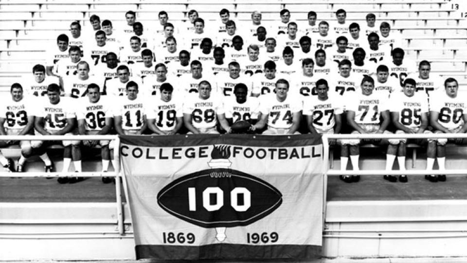 football team members in uniform posed on bleachers, with a sign reading "College Football 100 / 1869-1969" unfurled in front
