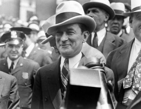 The charming, dapper, witty and utterly corrupt Jimmy Walker, mayor of New York from 1926 to 1932. Man in a fedora smiles while posed in front of a large group of men with a policeman looking on