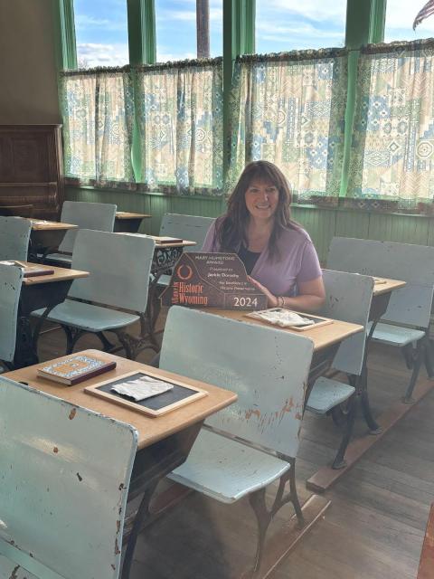 A smiling woman holding an award sits at a wooden student desk in an old schoolhouse