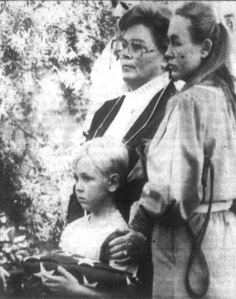 Two women stand behind a young boy who is holding a folded flag.