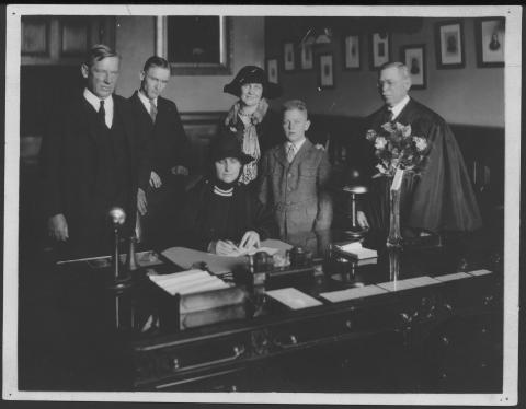 Three men, a woman and a boy in formal dress pose, standing behind a woman seated at a desk. She looks down, holding pen to paper.