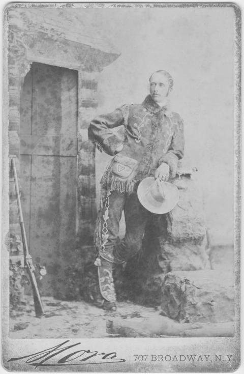 Man dressed in buckskins holding a cowboy hat poses next to a doorway