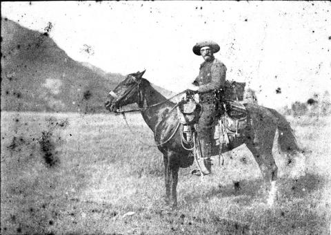 man in cowboy hat sits astride a Western saddle on a horse loaded with gear