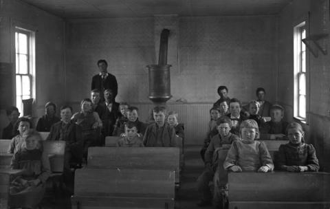 Formal portrait of a schoolroom, students of varying ages seated at desks with the teacher at the back of the room standing near a woodstove