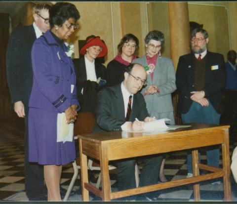 A group of people standing behind Wyo. Gov. Mike Sullivan watch him sign some papers 