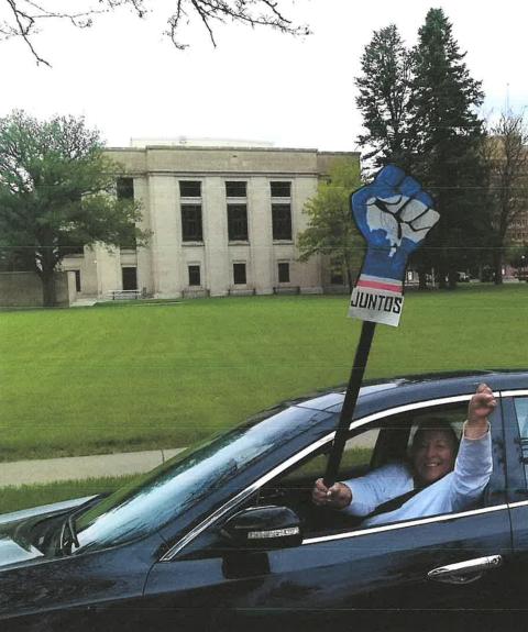 A woman in the driver's seat of a car holds a poster of a clenched fist with the word, "Juntos," out the window, while raising her left hand in a clenched fist